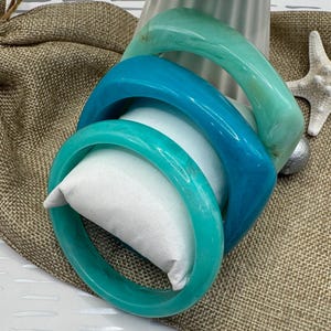 May include: Four colorful bangles in shades of teal and turquoise. The bangles are stacked on a white pillow, with a burlap background. The bangles have a glossy finish and a smooth texture. The bangles are a fashionable accessory.
