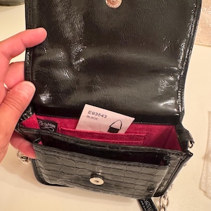 Op de afbeelding: Een zwarte crossbodytas van lakleer met een krokodil-reli&euml;f, voorzien van een magnetische drukknoopsluiting, een felroze binnenvoering en een zilverkleurige kettingriem. Een wit label aan de binnenzijde toont de tekst E93543 BLACK.