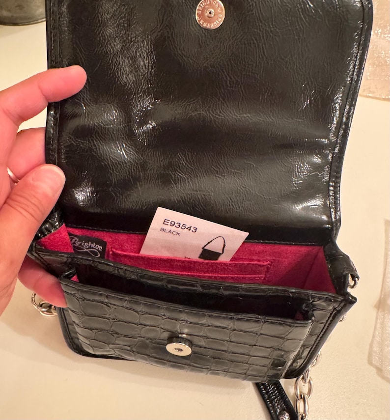 Op de afbeelding: Een zwarte crossbodytas van lakleer met een krokodil-reli&euml;f, voorzien van een magnetische drukknoopsluiting, een felroze binnenvoering en een zilverkleurige kettingriem. Een wit label aan de binnenzijde toont de tekst E93543 BLACK.
