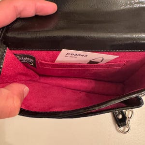 Op de afbeelding: Een zwarte handtas met een glanzende buitenkant en een felroze binnenvoering. De tas is voorzien van een Brighton-merklabel en een witte productinformatiekaart met de tekst E93543 BLACK, die in een binnenvak is gestoken.