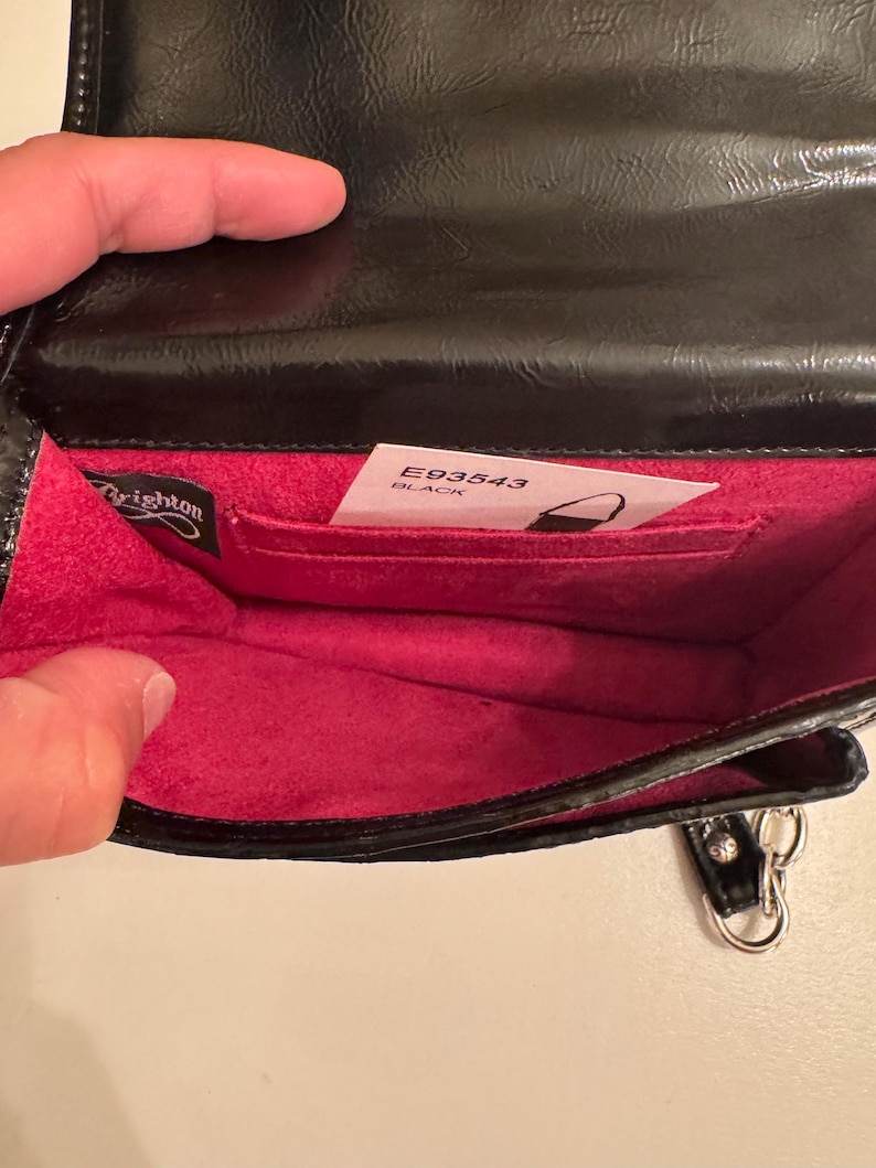 Op de afbeelding: Een zwarte handtas met een glanzende buitenkant en een felroze binnenvoering. De tas is voorzien van een Brighton-merklabel en een witte productinformatiekaart met de tekst E93543 BLACK, die in een binnenvak is gestoken.