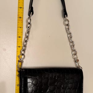 Op de afbeelding: Een zwarte handtas met een glanzende krokodil-reli&euml;ftextuur, voorzien van een zilverkleurige metalen kettinghengsel en een decoratieve ronde zilveren knoop op de voorflap. Een geel meetlint toont een hoogte van ongeveer 28 cm.