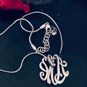 Op de afbeelding: Zilverkleurige ketting met een monogram hanger. De hanger heeft een sierlijk ontwerp van de letters "JM". De ketting heeft een fijne, verstelbare ketting met een karabijnsluiting. De achtergrond is donker en effen.