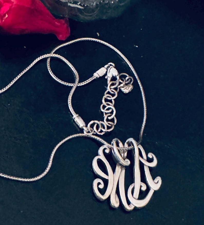 Op de afbeelding: Zilverkleurige ketting met een monogram hanger. De hanger heeft een sierlijk ontwerp van de letters "JM". De ketting heeft een fijne, verstelbare ketting met een karabijnsluiting. De achtergrond is donker en effen.