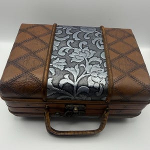 May include: A rectangular storage case featuring brown faux leather sides with a diamond-stitched pattern and a central vertical panel of silver floral fabric. The case includes a brown handle and a metal latch closure on the front edge.