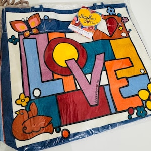 Op de afbeelding: Een witte draagtas met een kleurrijk "LOVE"-ontwerp. Het woord "LOVE" staat in grote blokletters met een gele cirkel in de "O". De tas heeft een blauwe rand en heeft een vlinder, vogel en bloemen. Een label leest "Brighton" en "Power of Love Tote."