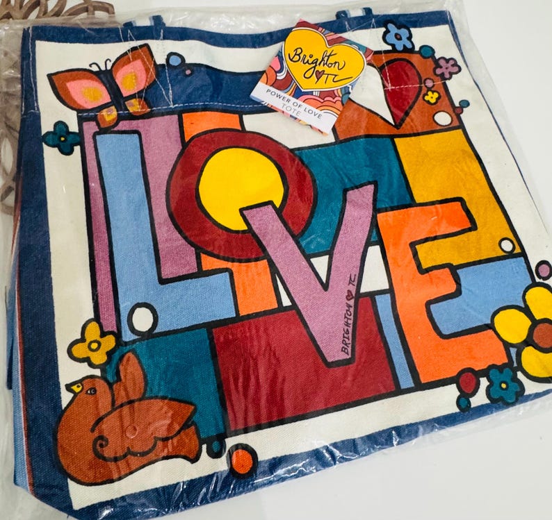 Op de afbeelding: Een witte draagtas met een kleurrijk "LOVE"-ontwerp. Het woord "LOVE" staat in grote blokletters met een gele cirkel in de "O". De tas heeft een blauwe rand en heeft een vlinder, vogel en bloemen. Een label leest "Brighton" en "Power of Love Tote."