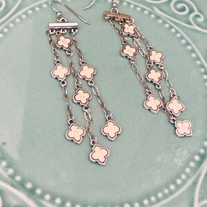 May include: A pair of silver-toned dangle earrings featuring three delicate chains suspended from a horizontal bar. Each chain is adorned with multiple small, cream-colored quatrefoil-shaped charms with silver-toned metal borders and hook wires.