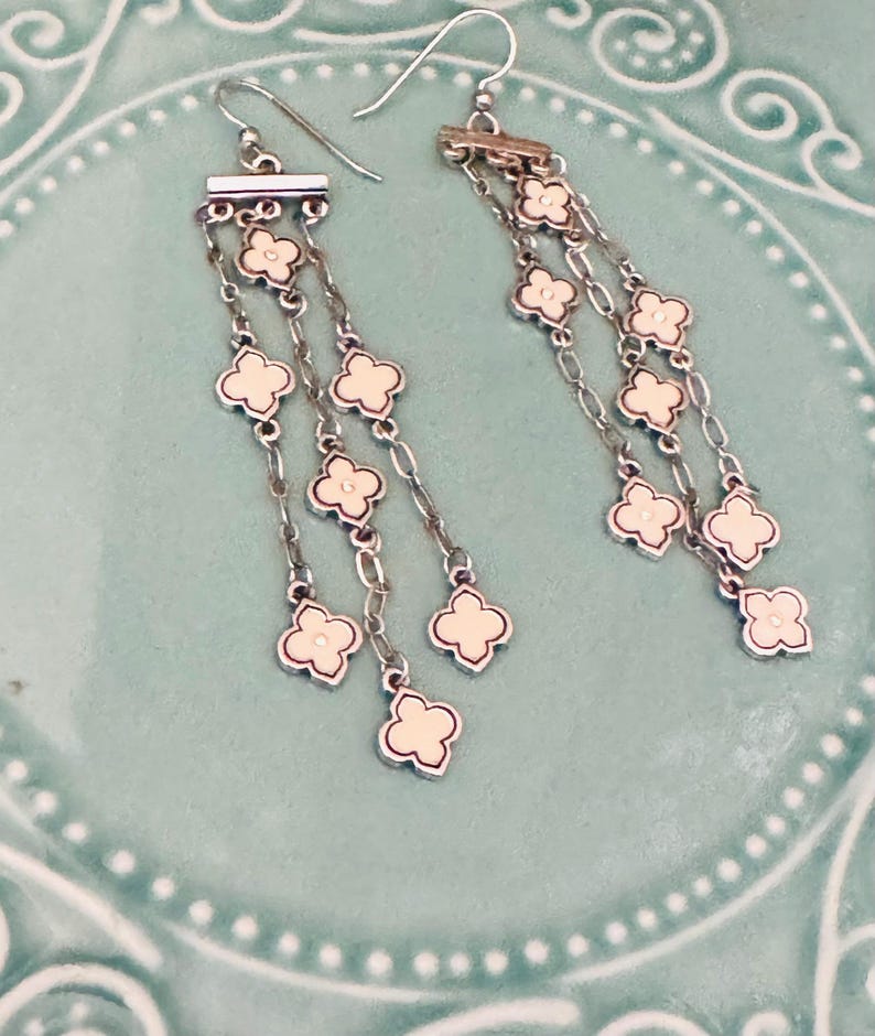May include: A pair of silver-toned dangle earrings featuring three delicate chains suspended from a horizontal bar. Each chain is adorned with multiple small, cream-colored quatrefoil-shaped charms with silver-toned metal borders and hook wires.