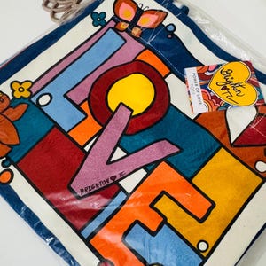 Op de afbeelding: Een kleurrijke draagtas met het woord "LOVE" in vette letters. Het ontwerp heeft een geometrisch patroon met levendige kleuren zoals rood, oranje, blauw en geel. Een label leest "Brighton Tote".