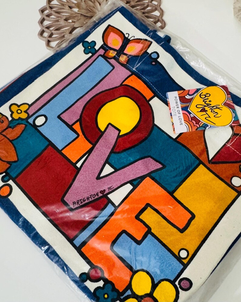 Op de afbeelding: Een kleurrijke draagtas met het woord "LOVE" in vette letters. Het ontwerp heeft een geometrisch patroon met levendige kleuren zoals rood, oranje, blauw en geel. Een label leest "Brighton Tote".