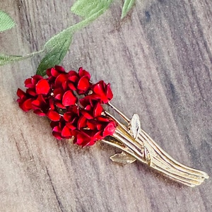 May include: A gold-toned metal brooch in the shape of a bouquet of red flowers. The flowers are clustered together, with individual petals visible. The stems are gold and have small leaf accents.