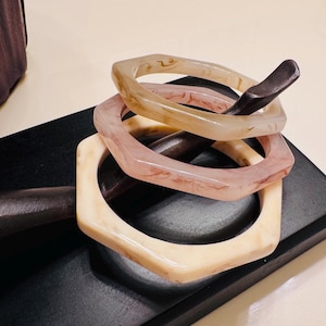 May include: Three hexagonal bangles in shades of cream, pink, and tan. The bangles are stacked on a dark, rectangular surface. The bracelets have a marbled appearance, with subtle variations in color and texture. The overall aesthetic is modern and minimalist.