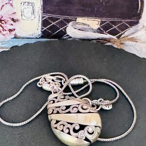 May include: Silver-toned pendant necklace with an oval, filigree-detailed charm. The charm has a central band and intricate scrollwork. The necklace is displayed on a dark surface, with a perfume bottle and a black handbag in the background.