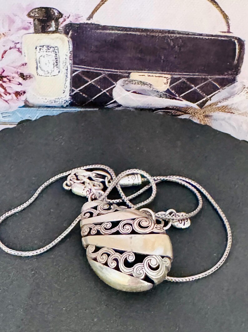 May include: Silver-toned pendant necklace with an oval, filigree-detailed charm. The charm has a central band and intricate scrollwork. The necklace is displayed on a dark surface, with a perfume bottle and a black handbag in the background.