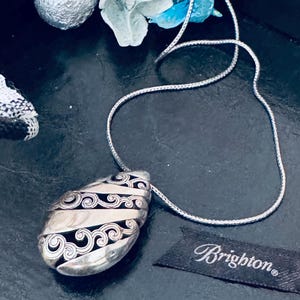 May include: Silver-toned pendant necklace with a teardrop-shaped pendant. The pendant features a swirling, patterned design with black accents. The necklace has a delicate chain and the Brighton brand tag is visible.