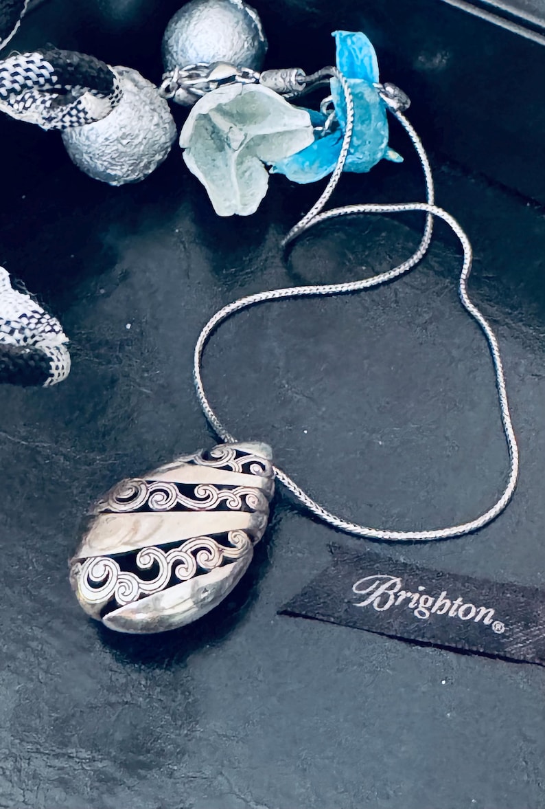 May include: Silver-toned pendant necklace with a teardrop-shaped pendant. The pendant features a swirling, patterned design with black accents. The necklace has a delicate chain and the Brighton brand tag is visible.