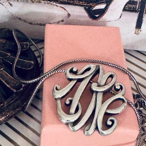 Op de afbeelding: Zilverkleurige monogramketting met een fijne ketting, op een roze doos. Het monogram toont verweven letters. De achtergrond bevat een decoratieve print met het woord "PARIS" en een pumps.