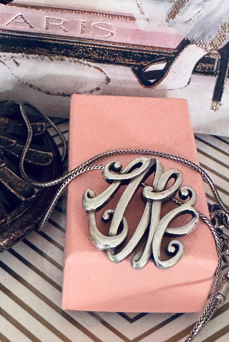 Op de afbeelding: Zilverkleurige monogramketting met een fijne ketting, op een roze doos. Het monogram toont verweven letters. De achtergrond bevat een decoratieve print met het woord "PARIS" en een pumps.