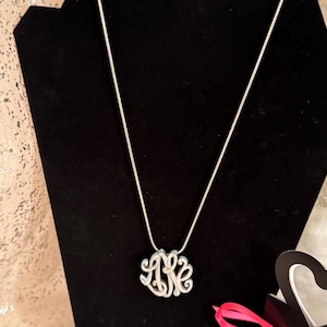 Op de afbeelding: Een zilverkleurige monogramketting met een fijne ketting wordt getoond op een zwarte fluwelen buste. Het monogram bevat de letters L, K en E in een gestileerd, verweven ontwerp. De ketting is een klassiek sieraad.