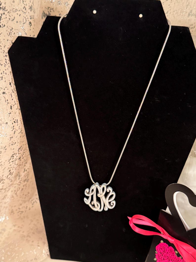 Op de afbeelding: Een zilverkleurige monogramketting met een fijne ketting wordt getoond op een zwarte fluwelen buste. Het monogram bevat de letters L, K en E in een gestileerd, verweven ontwerp. De ketting is een klassiek sieraad.