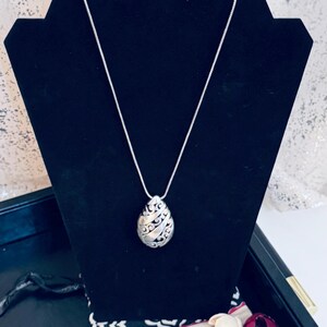 May include: Silver-toned pendant necklace with an oval, openwork design. The pendant hangs from a delicate chain and is displayed on a black jewelry stand. A black and white patterned cloth and a tag with the word "Brighton" are in the background.