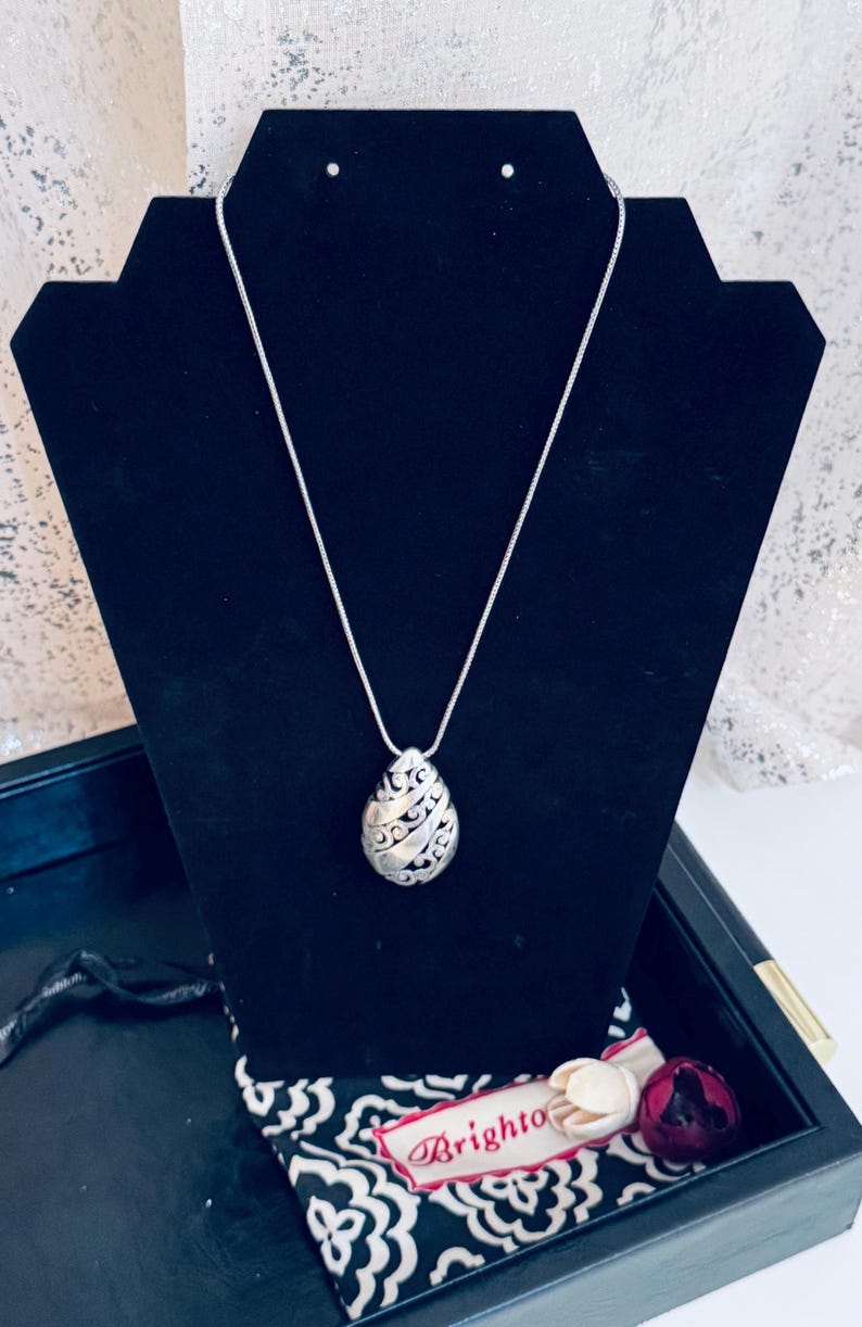 May include: Silver-toned pendant necklace with an oval, openwork design. The pendant hangs from a delicate chain and is displayed on a black jewelry stand. A black and white patterned cloth and a tag with the word "Brighton" are in the background.