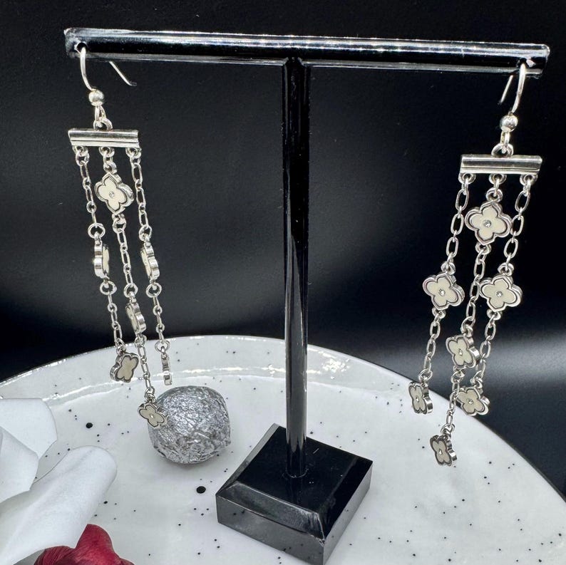 May include: A pair of silver-toned dangle earrings featuring three cascading chains suspended from a horizontal bar. Each chain is adorned with multiple small, four-petaled clover-shaped charms with cream-colored centers and silver edges.