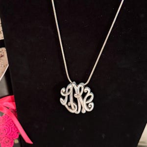 Op de afbeelding: Een zilverkleurige monogramketting met een fijne ketting. De hanger heeft een verstrengeld ontwerp van drie letters, waarschijnlijk initialen, in een cursieve stijl. De ketting wordt gepresenteerd tegen een zwarte achtergrond, waardoor de metallic glans wordt benadrukt.