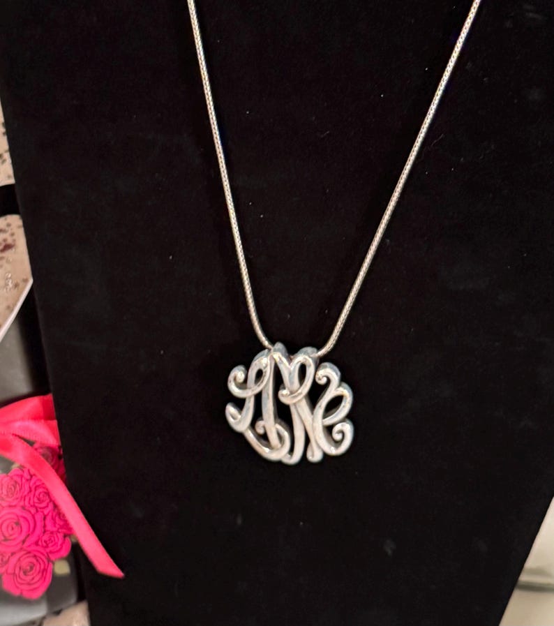 Op de afbeelding: Een zilverkleurige monogramketting met een fijne ketting. De hanger heeft een verstrengeld ontwerp van drie letters, waarschijnlijk initialen, in een cursieve stijl. De ketting wordt gepresenteerd tegen een zwarte achtergrond, waardoor de metallic glans wordt benadrukt.