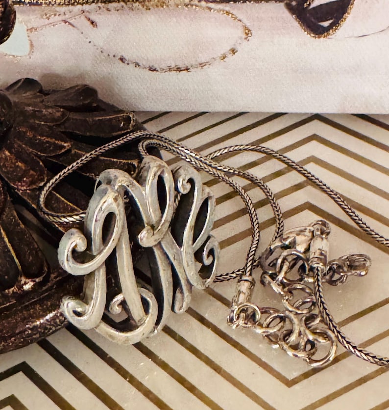 Op de afbeelding: Zilveren monogramketting met een gedetailleerd, wervelend ontwerp. De hanger heeft de verweven letters "MNR" en hangt aan een delicate ketting. De ketting wordt gepresenteerd op een patroonoppervlak.