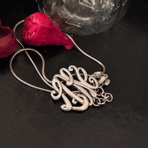 Op de afbeelding: Een zilveren ketting met een sierlijke monogramhanger. De hanger heeft verweven letters in een vloeiend schrift. De ketting ligt op een zwart oppervlak, met een rood bloemblad en een glazen fles op de achtergrond.