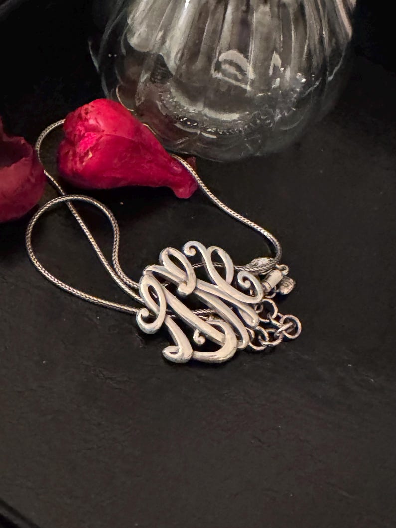 Op de afbeelding: Een zilveren ketting met een sierlijke monogramhanger. De hanger heeft verweven letters in een vloeiend schrift. De ketting ligt op een zwart oppervlak, met een rood bloemblad en een glazen fles op de achtergrond.