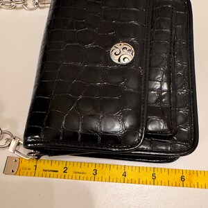 Op de afbeelding: Een zwarte rechthoekige portemonnee met een glanzende krokodil-reli&euml;ftextuur en een zilverkleurige ronde decoratieve knop. Een geel meetlint ligt langs de onderrand en toont een breedte van ongeveer 16,5 cm.