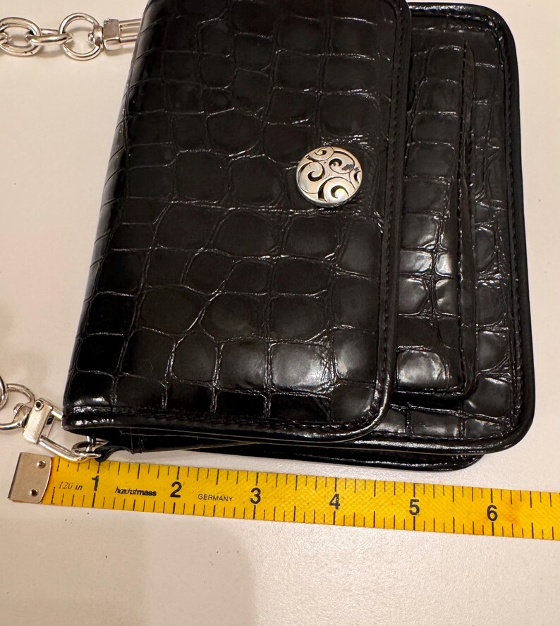 Op de afbeelding: Een zwarte rechthoekige portemonnee met een glanzende krokodil-reli&euml;ftextuur en een zilverkleurige ronde decoratieve knop. Een geel meetlint ligt langs de onderrand en toont een breedte van ongeveer 16,5 cm.