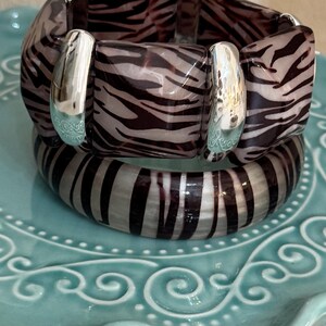 Puede incluir: Dos pulseras apiladas con un estampado de cebra en tonos marrón y crema. La pulsera superior tiene eslabones cuadrados y gruesos con conectores plateados. La pulsera inferior tiene un diseño redondeado. Las pulseras se exhiben en un plato decorativo azul claro.