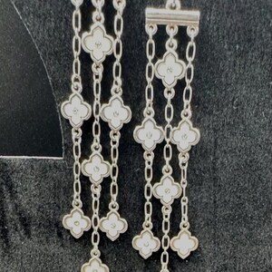May include: A pair of silver-toned drop earrings featuring three vertical chains suspended from a horizontal bar. Each chain is adorned with multiple white four-petaled clover-shaped charms, each containing a small central dot detail.