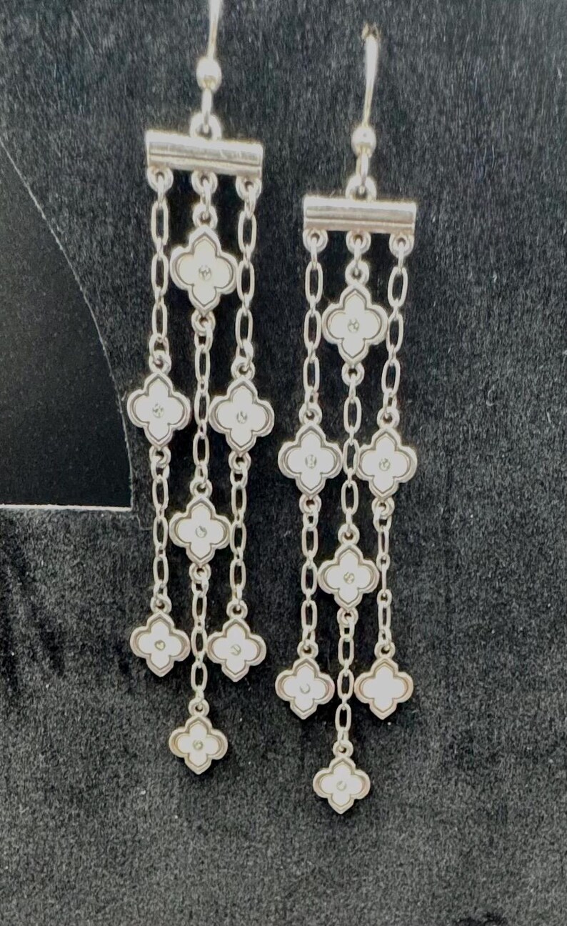 May include: A pair of silver-toned drop earrings featuring three vertical chains suspended from a horizontal bar. Each chain is adorned with multiple white four-petaled clover-shaped charms, each containing a small central dot detail.