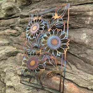 May include: Metal sunflower wall art with a cut-out design. The sunflowers have a gradient of colors, including bronze, blue, and gold. The artwork is rectangular and features three large sunflowers and several smaller buds and leaves.