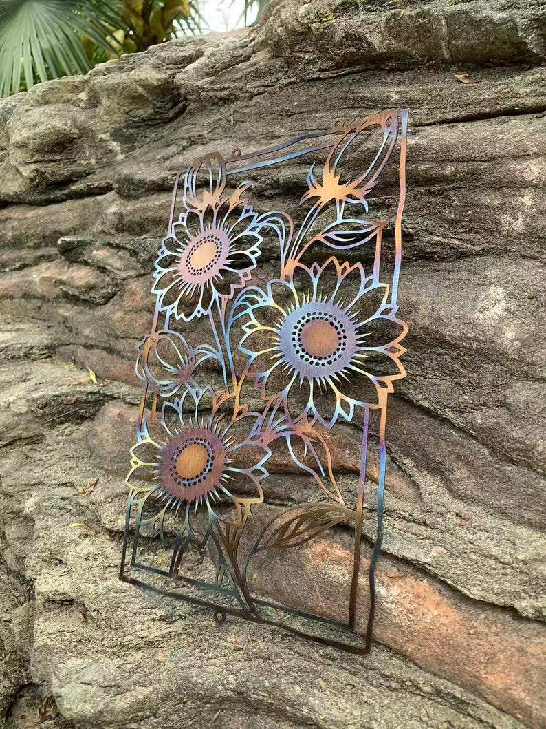 May include: Metal sunflower wall art with a cut-out design. The sunflowers have a gradient of colors, including bronze, blue, and gold. The artwork is rectangular and features three large sunflowers and several smaller buds and leaves.