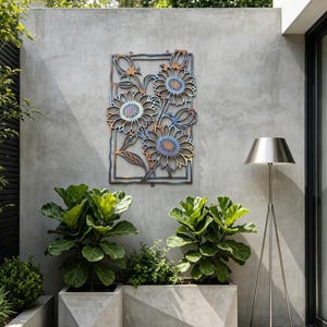 May include: Metal wall art featuring a floral design with sunflowers in shades of blue, orange, and brown. The artwork is mounted on a gray concrete wall in an outdoor setting. The art is framed with a rectangular border.