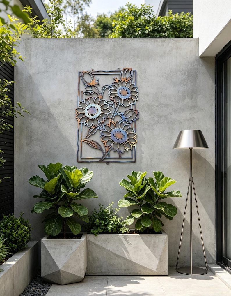 May include: Metal wall art featuring a floral design with sunflowers in shades of blue, orange, and brown. The artwork is mounted on a gray concrete wall in an outdoor setting. The art is framed with a rectangular border.