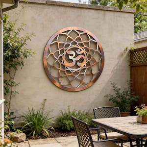 Pode incluir: Uma grande peça de arte de parede redonda de metal cor de cobre com um símbolo Om no centro. A arte tem um design intrincado e vazado e é montada em uma parede de estuque bege em um ambiente externo. A arte é cercada por vegetação.