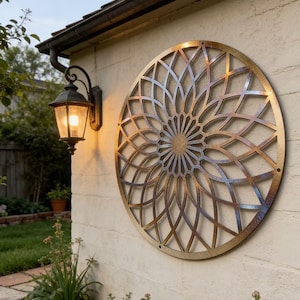 Op de afbeelding: Een groot, cirkelvormig metalen wandkunstwerk met een bloemenontwerp is bevestigd aan een getextureerde, gebroken witte muur. De kunst heeft een bronzen en blauwe afwerking. Links van het kunstwerk is een buitenlamp in vintage stijl gemonteerd, die een warme gloed uitstraalt.