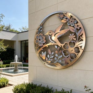 May include: Circular metal wall art depicting a hummingbird and floral design. The hummingbird sits on a branch surrounded by flowers and leaves. The artwork is mounted on a beige stone wall. A fountain and greenery are visible in the background.