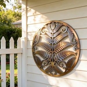 Op de afbeelding: Ronde metalen wandkunst met een varenontwerp. Het kunstwerk heeft brons-, zilver- en goudtinten, met ingewikkelde uitgesneden details. Het is gemonteerd op een witte muur naast een witte hek.