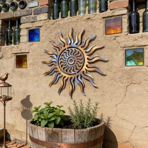 Könnte beinhalten: Eine dekorative Metallsonnenskulptur mit einem Wirbelzentrum und strahlenden Strahlen ist an einer strukturierten Wand angebracht. Die Wand weist Glasquadrate und wiederverwendete Flaschen auf. Darunter befindet sich ein Holzfass-Pflanzgefäß mit Grün.