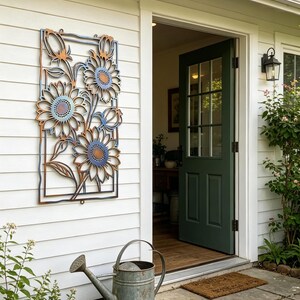 May include: A metal sunflower wall art piece with a rectangular frame is mounted on a white wall. The sunflowers have blue and brown accents. A galvanized metal watering can sits on a stone patio. A green door is open.