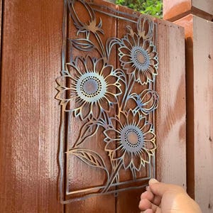 May include: A metal sunflower wall art piece with a rectangular frame. The art features three sunflowers with detailed petals and a central design. The metal has a gradient color effect, and the art is mounted on a brown wooden fence.
