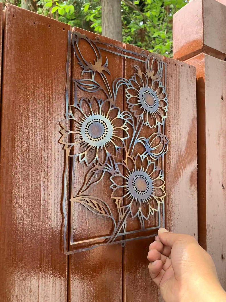 May include: A metal sunflower wall art piece with a rectangular frame. The art features three sunflowers with detailed petals and a central design. The metal has a gradient color effect, and the art is mounted on a brown wooden fence.